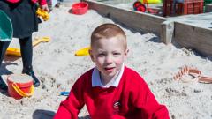 Young boy sat in the sandpit smiling for the camera early years playground