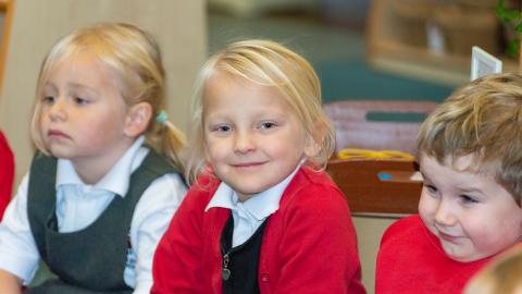 Early years pupils listening to the teacher, young girl smiles at the camera